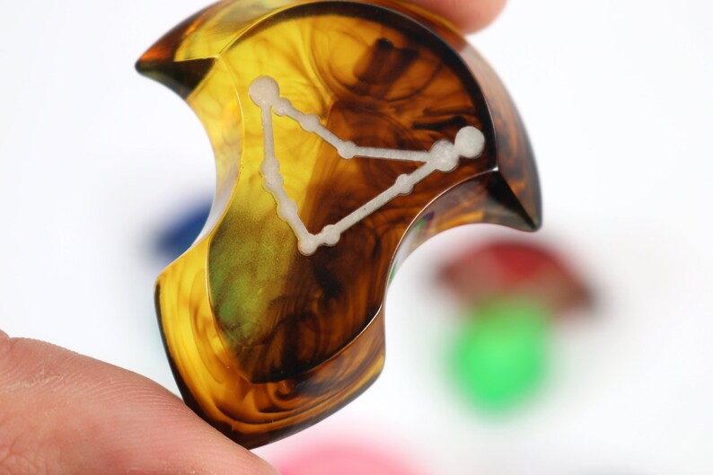 May include: A crescent-shaped, amber-coloured resin guitar pick with a white constellation design. The constellation is Pisces, the fish.