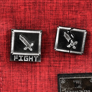 May include: Two silver metal pins with a pixelated sword design. The pin on the left says "FIGHT" below the sword. The pin on the right is broken in half.