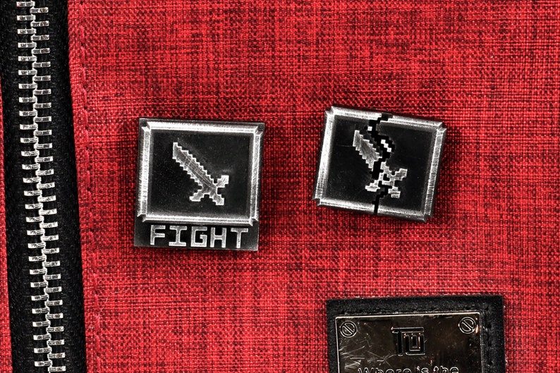 May include: Two silver metal pins with a pixelated sword design. The pin on the left says "FIGHT" below the sword. The pin on the right is broken in half.