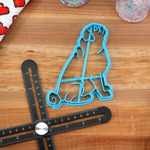 May include: Blue plastic cookie cutter shaped like a sitting pug dog. The cutter is on a wooden surface with a ruler and other tools.