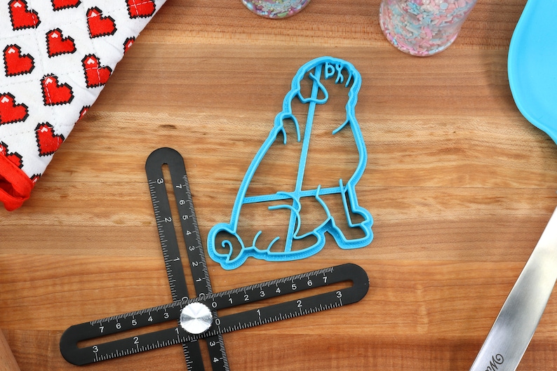 May include: Blue plastic cookie cutter shaped like a sitting pug dog. The cutter is on a wooden surface with a ruler and other tools.