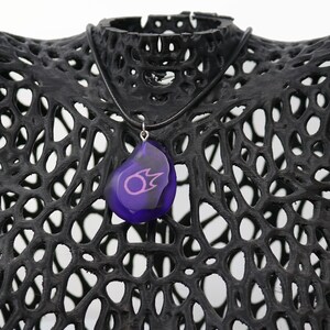 May include: A purple pendant necklace with a black cord. The pendant is shaped like a teardrop and has a pink symbol inside.