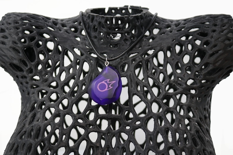 May include: A purple pendant necklace with a black cord. The pendant is shaped like a teardrop and has a pink symbol inside.