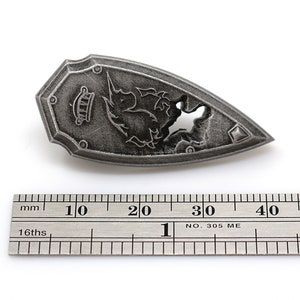 May include: A silver-colored shield-shaped pendant with a detailed design of a mythical creature. The pendant is approximately 3 cm long and is measured against a ruler.
