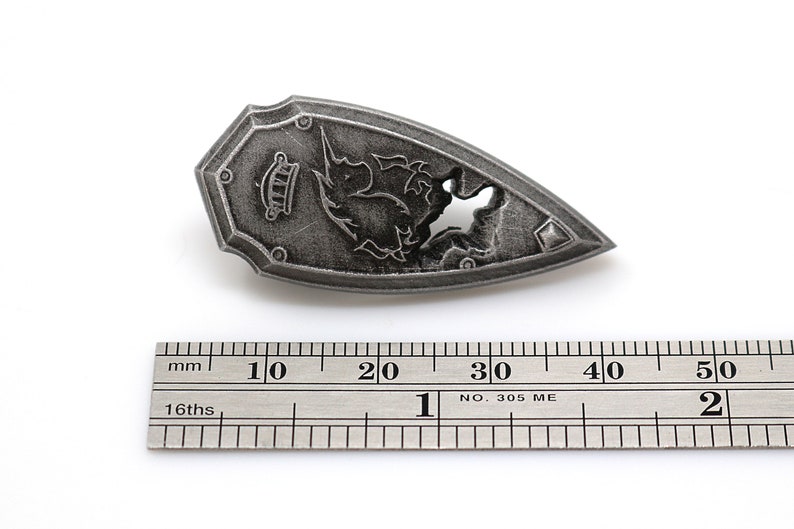 May include: A silver-colored shield-shaped pendant with a detailed design of a mythical creature. The pendant is approximately 3 cm long and is measured against a ruler.