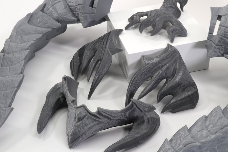 May include: A set of gray 3D printed dragon wings, claws, and tail pieces. The pieces are detailed with scales and spikes.