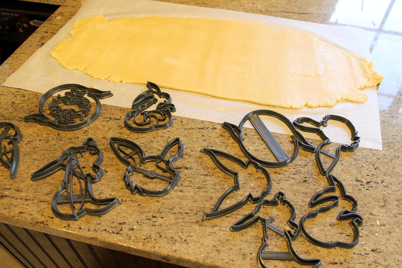 May include: A sheet of cookie dough on baking parchment with various cookie cutters in the shape of cartoon characters. The cookie cutters are made of black plastic.