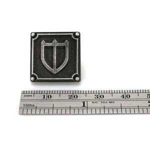 Puede incluir: Un pin de metal de color plateado con un dise&ntilde;o de escudo. El escudo est&aacute; bordeado de plata y tiene un fondo negro. El pin mide aproximadamente 2 cm de ancho.