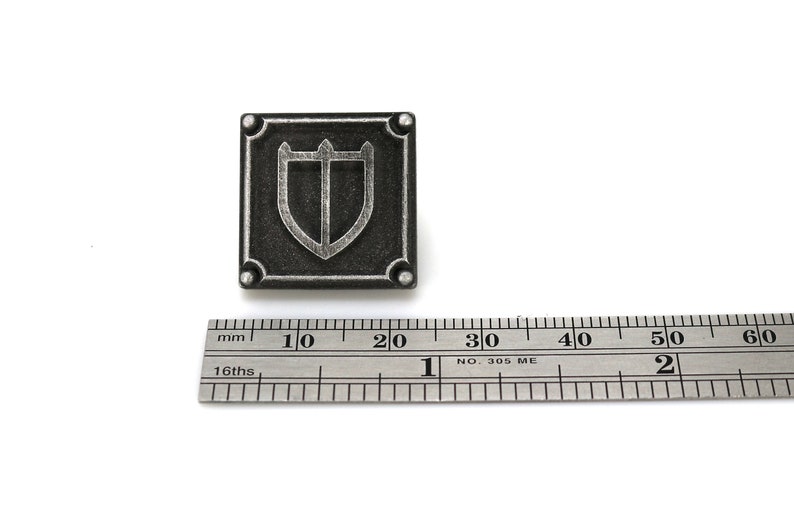 Puede incluir: Un pin de metal de color plateado con un dise&ntilde;o de escudo. El escudo est&aacute; bordeado de plata y tiene un fondo negro. El pin mide aproximadamente 2 cm de ancho.