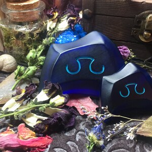 May include: Two blue glass dice with a symbol of a double-headed axe on each face. The dice are sitting on a dark surface with dried flowers and other objects.