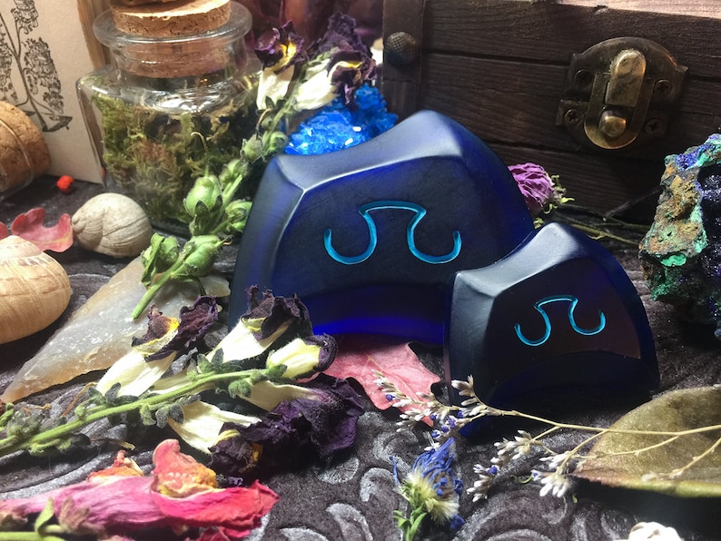 May include: Two blue glass dice with a symbol of a double-headed axe on each face. The dice are sitting on a dark surface with dried flowers and other objects.