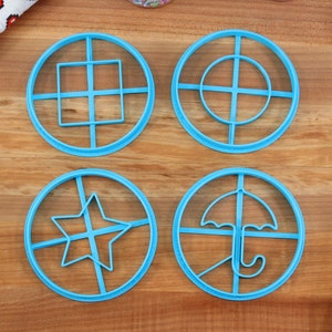 May include: Four light blue cookie cutters in circular shapes, each with a different design: a square grid, a circle with a crosshair, a star, and an umbrella. The cutters are on a wooden surface, with a patterned cloth and a spatula visible.