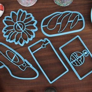 May include: A set of five blue plastic cookie cutters in various shapes, including a sunflower, a loaf of bread, a high-heeled shoe, and two rectangular shapes with a star and a circle design.