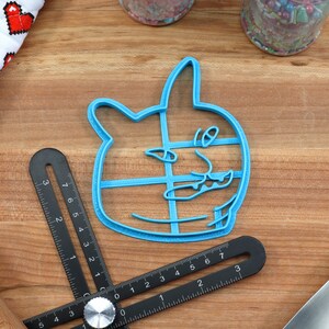 May include: A blue plastic cookie cutter shaped like a cat's face with a surprised expression. The cutter is on a wooden surface.