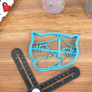 May include: A blue plastic cookie cutter shaped like a cat's face with large eyes and a surprised expression.