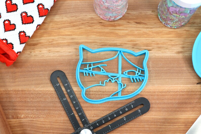 May include: A blue plastic cookie cutter shaped like a cat's face with large eyes and a surprised expression.