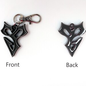 May include: A black metal keychain with a stylized design. The front of the keychain features a detailed, intricate design. The back of the keychain is smooth and has a silver ring.