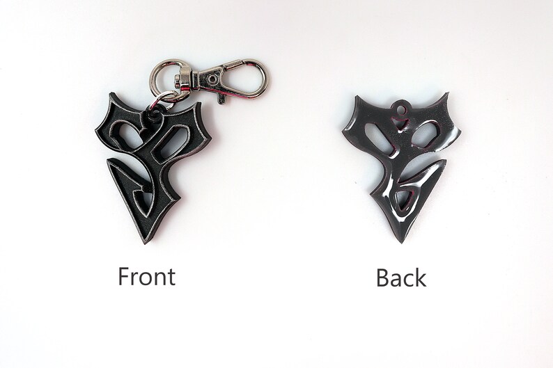 May include: A black metal keychain with a stylized design. The front of the keychain features a detailed, intricate design. The back of the keychain is smooth and has a silver ring.