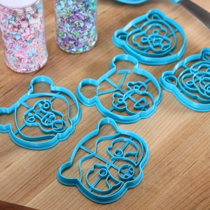 May include: Five blue plastic bear-shaped cookie cutters on a wooden surface. The cutters are different sizes and have intricate details.