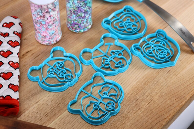 May include: Five blue plastic bear-shaped cookie cutters on a wooden surface. The cutters are different sizes and have intricate details.