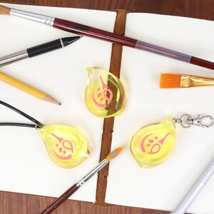May include: Three yellow teardrop-shaped charms with pink designs, one on a black cord necklace, one on a silver keychain, and one loose. The charms are on a white surface with pencils, pens, and paintbrushes.