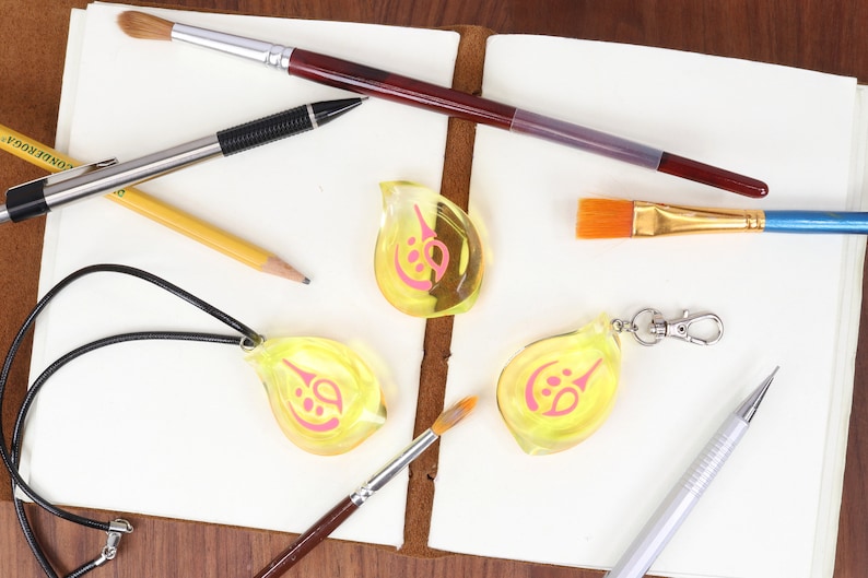 May include: Three yellow teardrop-shaped charms with pink designs, one on a black cord necklace, one on a silver keychain, and one loose. The charms are on a white surface with pencils, pens, and paintbrushes.