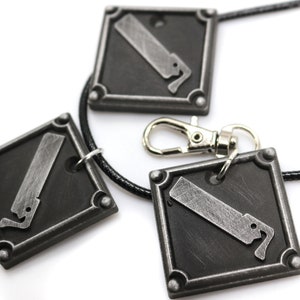 May include: Three silver-toned pendants with a saw blade design, attached to a black cord necklace. The pendants are square-shaped with rounded corners and have a textured surface.