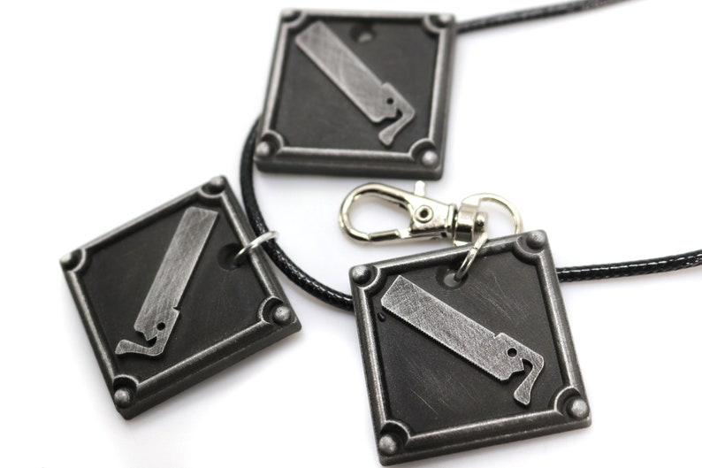 May include: Three silver-toned pendants with a saw blade design, attached to a black cord necklace. The pendants are square-shaped with rounded corners and have a textured surface.