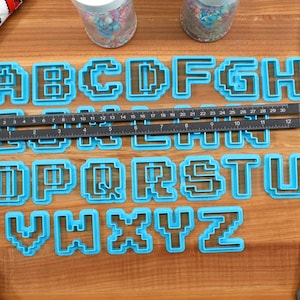 May include: A set of blue plastic alphabet cookie cutters in a pixelated font style. The cutters are arranged in alphabetical order on a wooden surface. A ruler and a spatula are also visible in the image.