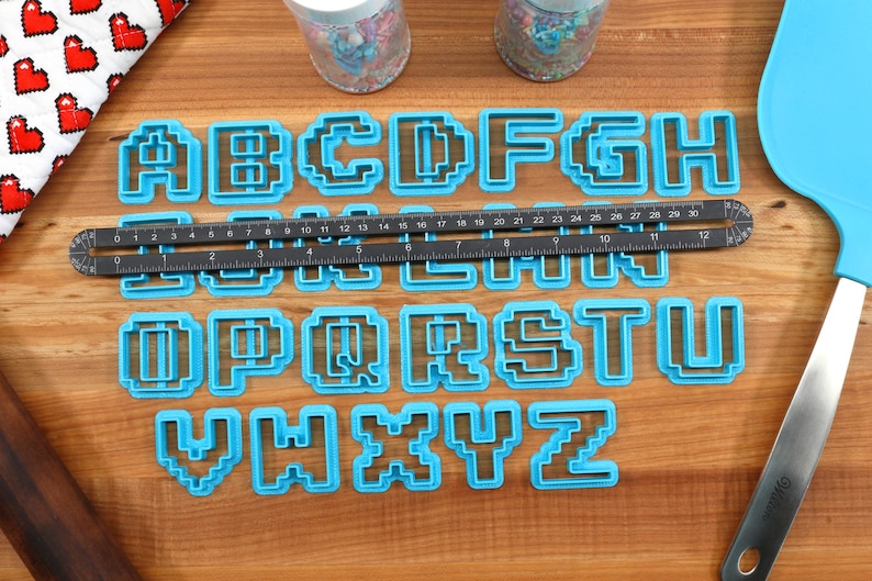 May include: A set of blue plastic alphabet cookie cutters in a pixelated font style. The cutters are arranged in alphabetical order on a wooden surface. A ruler and a spatula are also visible in the image.