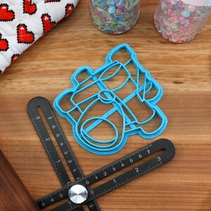 May include: A light blue cookie cutter in the shape of a baby carriage sits on a wooden surface. A black measuring tool, a rolling pin, and a blue spatula are also present. A white and red heart-patterned oven mitt is in the background.