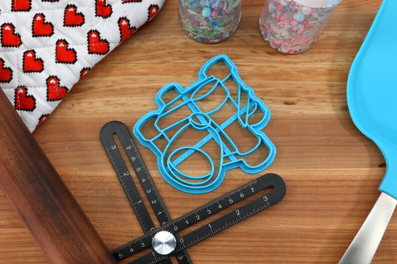 May include: A light blue cookie cutter in the shape of a baby carriage sits on a wooden surface. A black measuring tool, a rolling pin, and a blue spatula are also present. A white and red heart-patterned oven mitt is in the background.