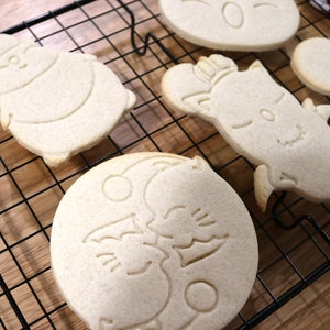 May include: Five sugar cookies on a black wire cooling rack. The cookies are decorated with various designs, including a snowman, a cat, a moon with two cats, and a heart.