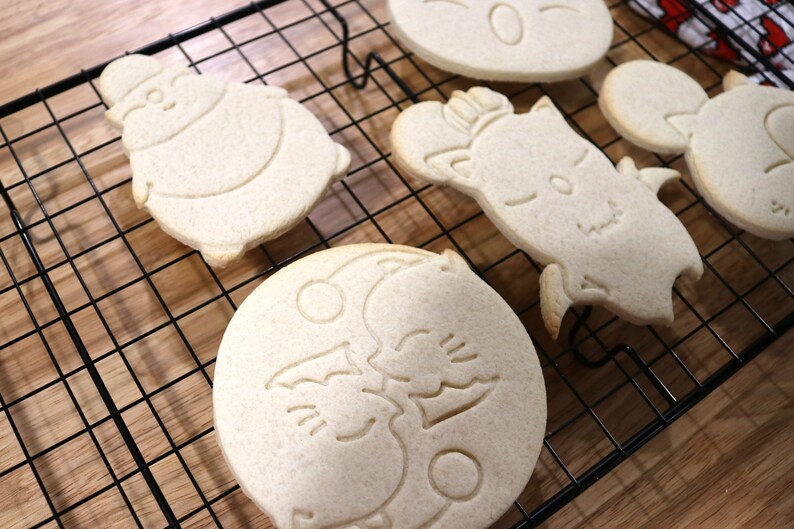 May include: Five sugar cookies on a black wire cooling rack. The cookies are decorated with various designs, including a snowman, a cat, a moon with two cats, and a heart.
