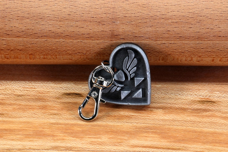 Puede incluir: Un llavero de coraz&oacute;n plateado con un s&iacute;mbolo alado y un dise&ntilde;o triangular. El llavero tiene un cierre de metal plateado.