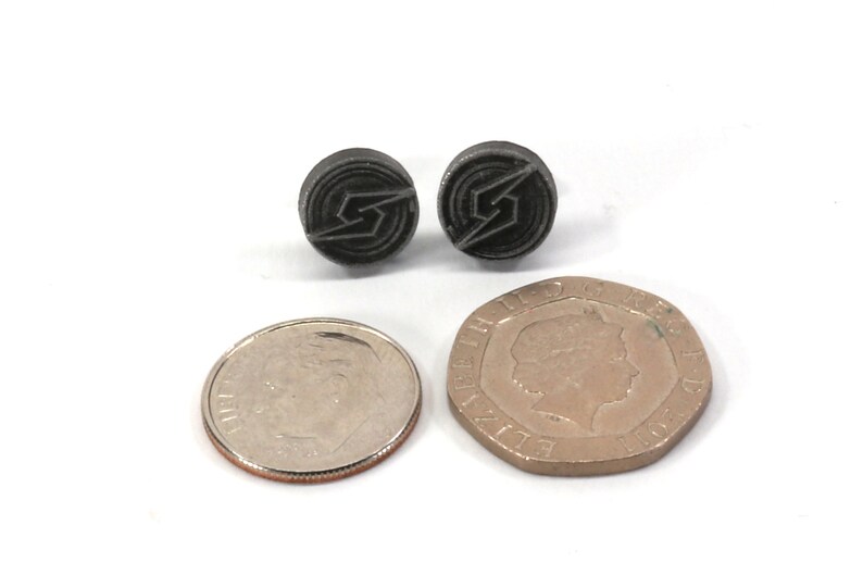 May include: A pair of black metal earrings featuring a stylised lightning bolt design. The earrings are shown next to a US dime and a British one pound coin for size comparison.