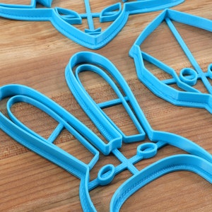 May include: A set of blue plastic cookie cutters in various shapes, including a bunny, a spaceship, and a geometric design. The cutters are on a wooden surface.