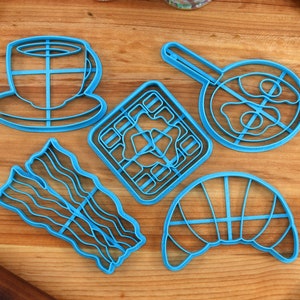May include: A collection of blue plastic cookie cutters in various breakfast-themed shapes, including a coffee cup, waffle, fried egg, bacon strips, and a croissant. The cutters are arranged on a wooden surface.