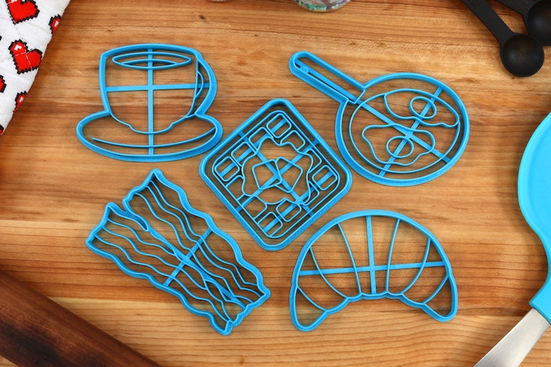 May include: A collection of blue plastic cookie cutters in various breakfast-themed shapes, including a coffee cup, waffle, fried egg, bacon strips, and a croissant. The cutters are arranged on a wooden surface.