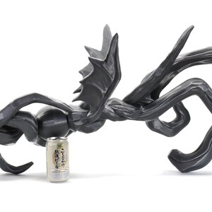 Puede incluir: Una escultura abstracta negra que se asemeja a un drag&oacute;n o una criatura m&iacute;tica con alas. La escultura est&aacute; hecha de un material liso y texturizado y tiene aproximadamente el tama&ntilde;o de una lata de refresco.