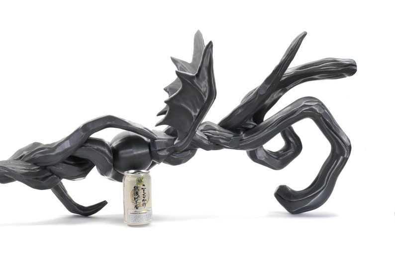 Puede incluir: Una escultura abstracta negra que se asemeja a un drag&oacute;n o una criatura m&iacute;tica con alas. La escultura est&aacute; hecha de un material liso y texturizado y tiene aproximadamente el tama&ntilde;o de una lata de refresco.