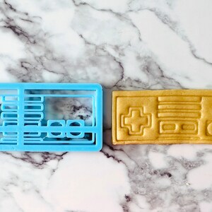 May include: A blue plastic cookie cutter and a light brown cookie in the shape of a vintage video game controller.