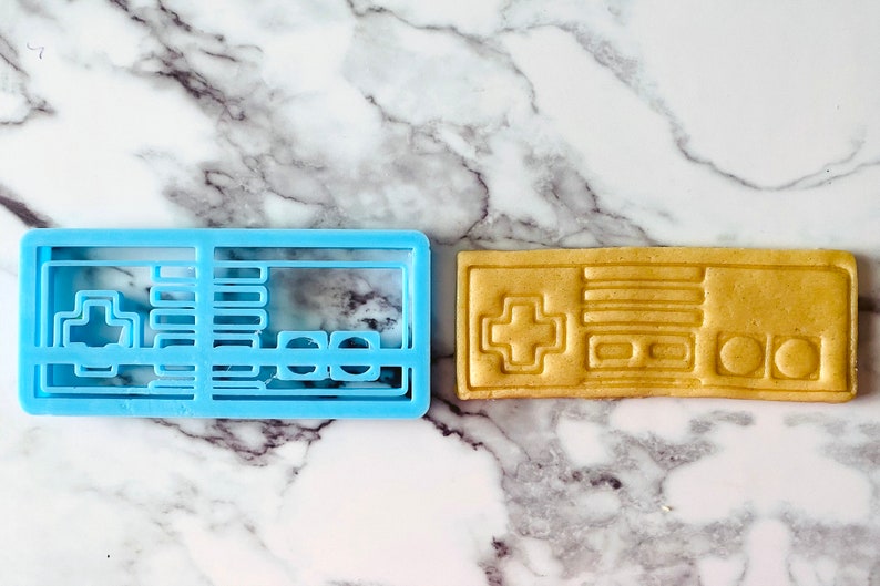 May include: A blue plastic cookie cutter and a light brown cookie in the shape of a vintage video game controller.