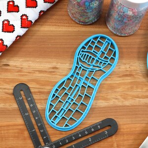 Puede incluir: Un cortador de galletas de pl&aacute;stico azul en forma de cacahuete con un patr&oacute;n de cuadr&iacute;cula. El cortador est&aacute; sobre una superficie de madera con una regla y una esp&aacute;tula.