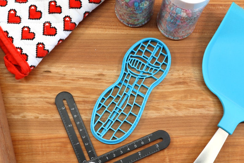 Puede incluir: Un cortador de galletas de pl&aacute;stico azul en forma de cacahuete con un patr&oacute;n de cuadr&iacute;cula. El cortador est&aacute; sobre una superficie de madera con una regla y una esp&aacute;tula.