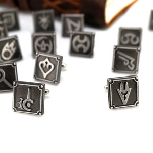 May include: A set of silver cufflinks featuring various symbols, including a sword, a shield, and a crescent moon. The cufflinks are scattered on a white surface.