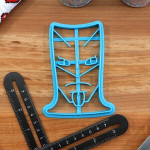 May include: A blue plastic cookie cutter shaped like a face with a tongue sticking out. The face has a serious expression with furrowed brows and a frown.