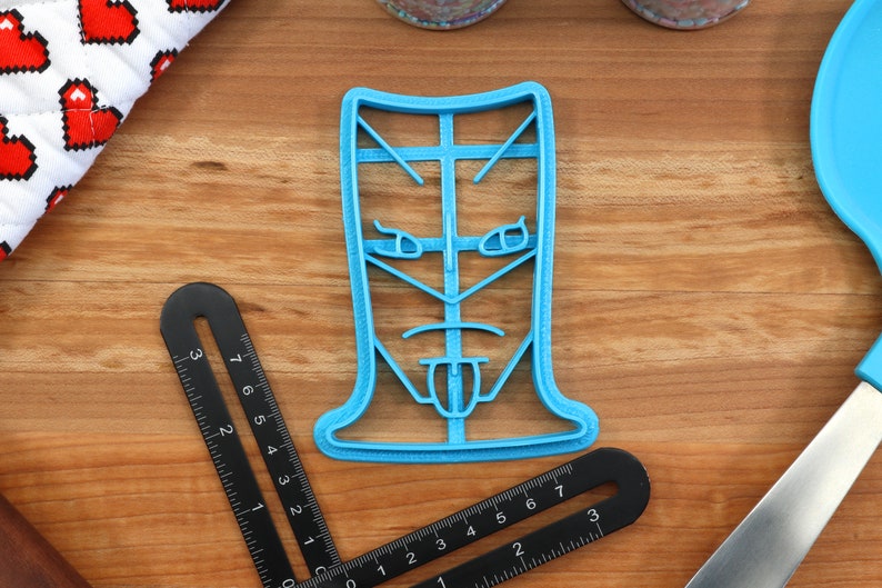 May include: A blue plastic cookie cutter shaped like a face with a tongue sticking out. The face has a serious expression with furrowed brows and a frown.