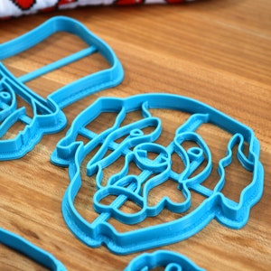 May include: Several light blue plastic cookie cutters in various shapes are arranged on a wooden surface. The cutters appear to be in the shape of animals and faces. A jar of sprinkles is visible in the background.