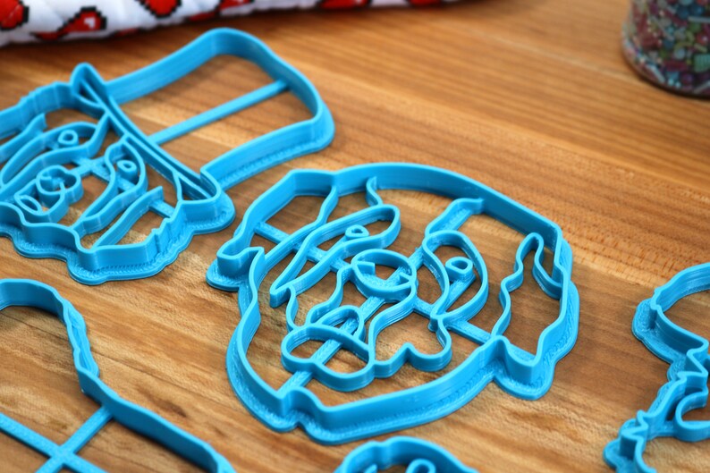 May include: Several light blue plastic cookie cutters in various shapes are arranged on a wooden surface. The cutters appear to be in the shape of animals and faces. A jar of sprinkles is visible in the background.